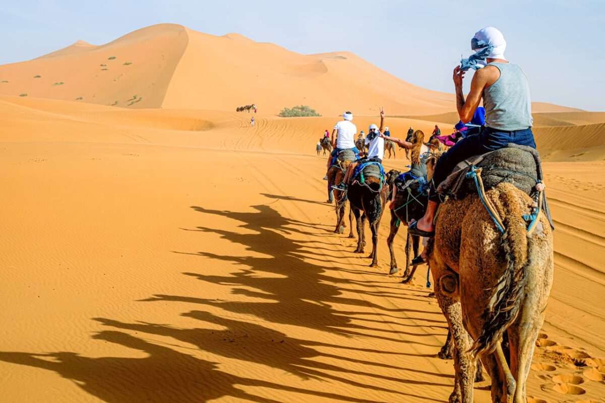 A group of people riding camels across the vast sand dunes under a bright sky.